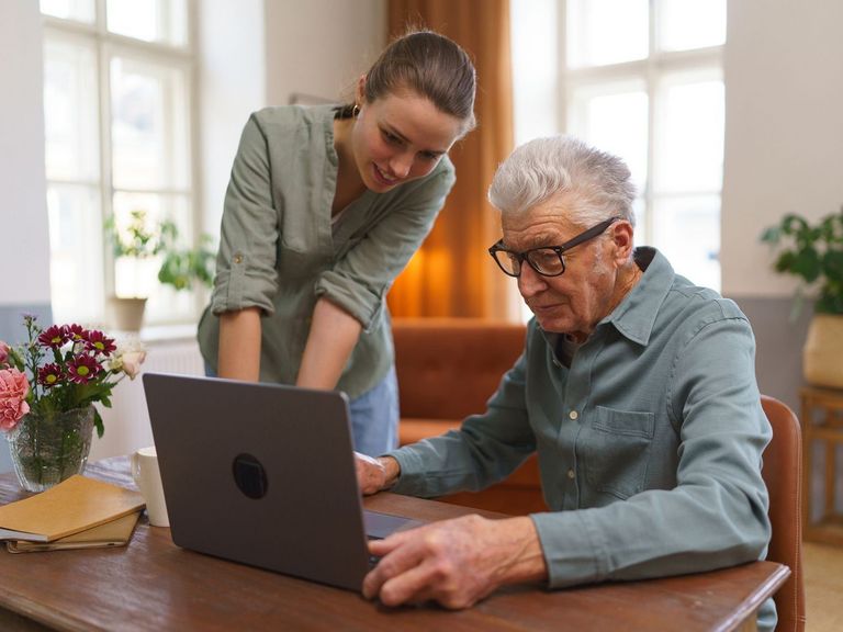 Rentner sitzt vor einem Laptop und erhält Hilfe von seiner erwachsenen Enkelin