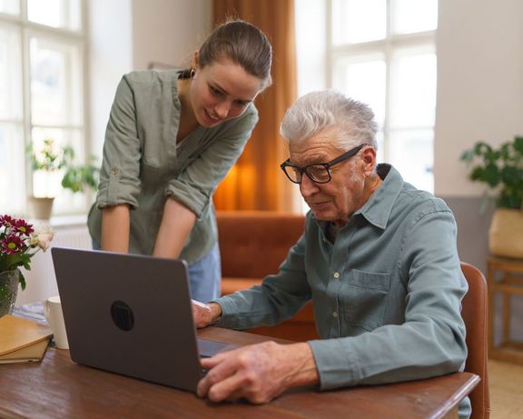 Rentner sitzt vor einem Laptop und erhält Hilfe von seiner erwachsenen Enkelin