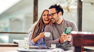 Digitaler Alltag Paar sitzt mit Smartphone und Kaffee lachend an einem Tisch