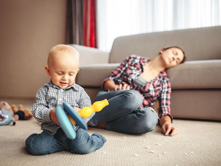 Baby spielt im Vordergrund und die Mutter sitzt erschöpft auf dem Boden und lehnt am Sofa