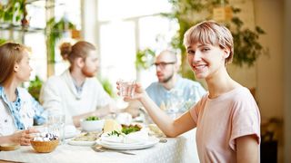 Trinken bei Mahlzeiten Vier Freunde sitzen zum gemeinsamen Essen an einem Tisch und einer der Frau schaut mit einem Wasserglas in der Hand in die Kamera