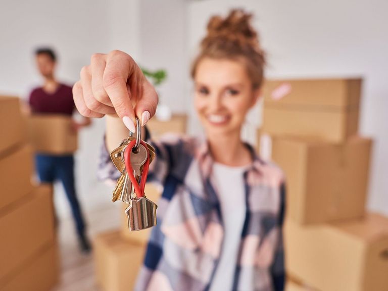 Junge Frau steht in einer Wohnung mit Umzugskartons und zeigt den Schlüssel direkt in die Kamera