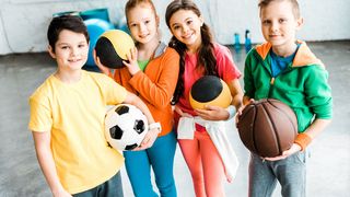 Kleiner Kindergruppe in Sportkleidung mit verschiedenen Ballarten in den Händen lacht in die Kamera