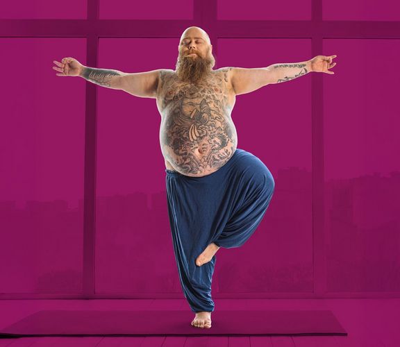 Bärtiger Mann mit Bauch und Tattoos steht auf einem Bein und streckt die Arme seitlich aus