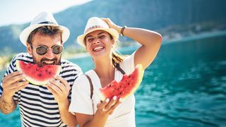 Essen im Sommer Mann und Frau vor einem See stehend, die lachend eine Melone essen