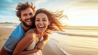 Junges Paar genießen lachend und mit Wind in den Haaren am Strand den Sonnenuntergang