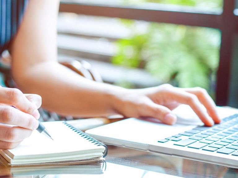 Frauenhände arbeiten an einem Laptop und daneben liegt ein Schreibblock