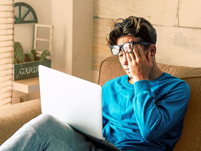 Jugendlicher sitzt auf der Couch, hat einen Laptop auf dem Schoß und reib sich mit einhändig die Augen und verschiebt dabei seine Brille