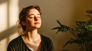 Junge Frau sitzt meditierend in der Sonne, die durch ein Fenster hereinscheint 