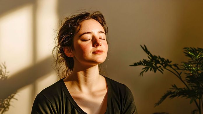 Junge Frau sitzt meditierend in der Sonne, die durch ein Fenster hereinscheint 