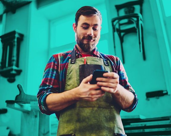 Handwerker in seiner Werkstatt schaut auf sein Smartphone