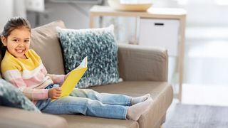 Geschichten zur Gesundheit Kleines Mädchen sitzt auf einer Couch und hat ein Buch in der Hand