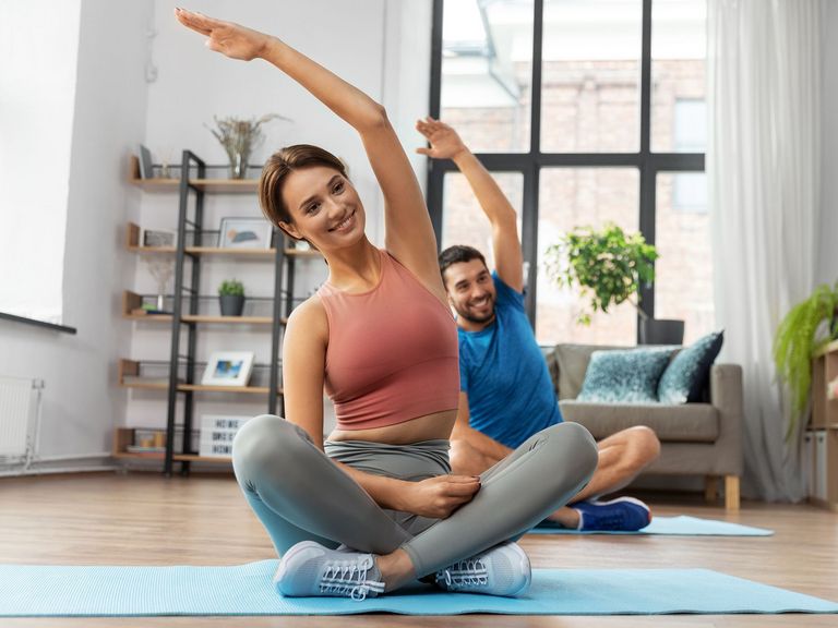 Pärchen im Wohnzimmer sitzt in Schneidersitz auf Yogamatten und macht Dehnübungen