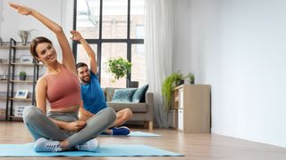 Bonusprogramm AchtzehnPlus Pärchen im Wohnzimmer sitzt in Schneidersitz auf Yogamatten und macht Dehnübungen