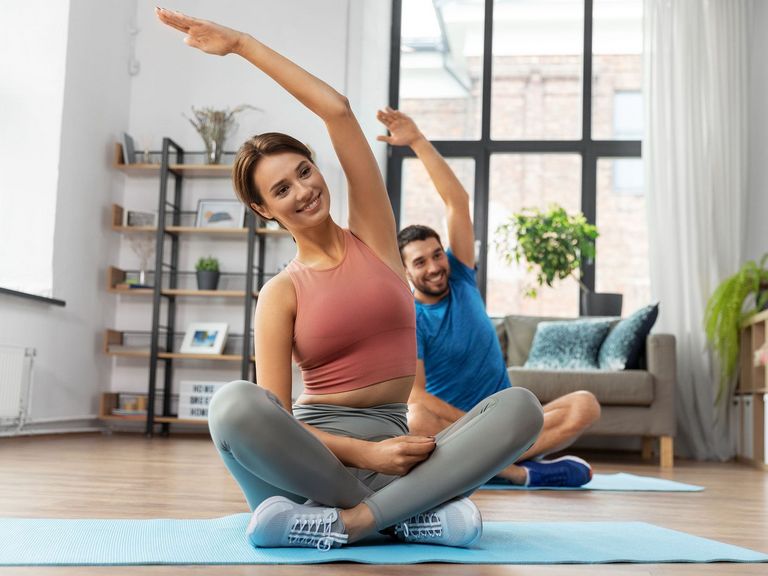 Pärchen im Wohnzimmer sitzt in Schneidersitz auf Yogamatten und macht Dehnübungen