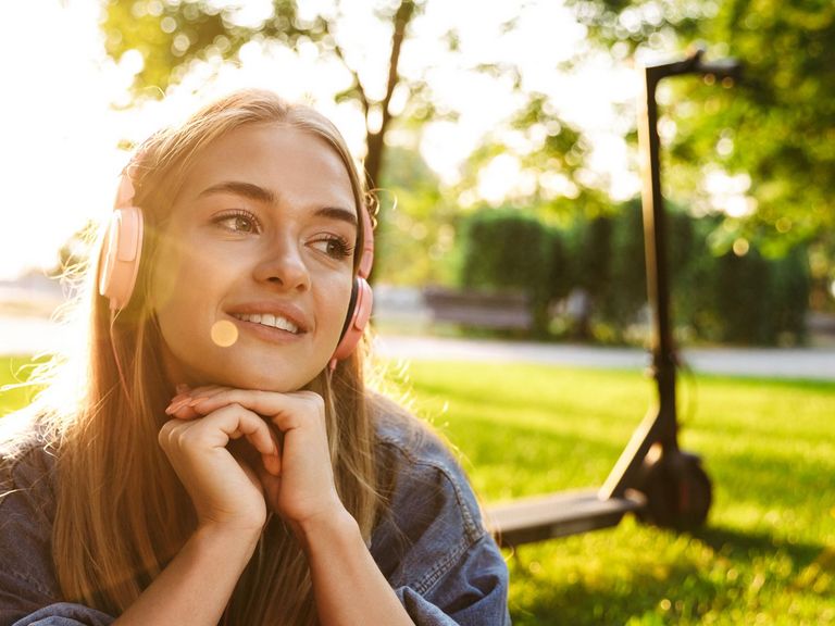 Junge Frau mit Kopfhörern sitzt im Park und hinter ihr steht ein E-Scooter