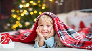Skandinavisches Hygge Kleiner, grinsender Junge mit einer karierten Decke auf dem Kopf und im Hintergrund leuchten Lichter