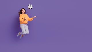 Mädchen im orangefarbenen Pullover springt mit einem Fußball in der Hand und vor einem lilafarbenen Hintergrund in die Luft
