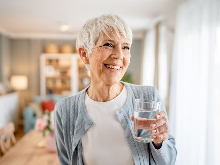 Frau lächelnd im Wohnzimmer mit einem Glas Wasser in der Hand
