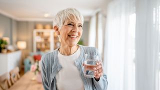 Frau lächelnd im Wohnzimmer mit einem Glas Wasser in der Hand