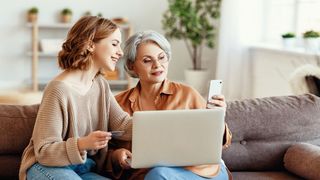 Zwei Frauen sitzen im Wohnzimmer auf einem Sofa und nutzen digitale Angebote per Laptop und Smartphone