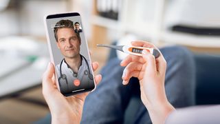 Hände halten ein Fieberthermometer und ein Smartphone, auf dem ein Videochat mit einem Arzt zu sehen ist