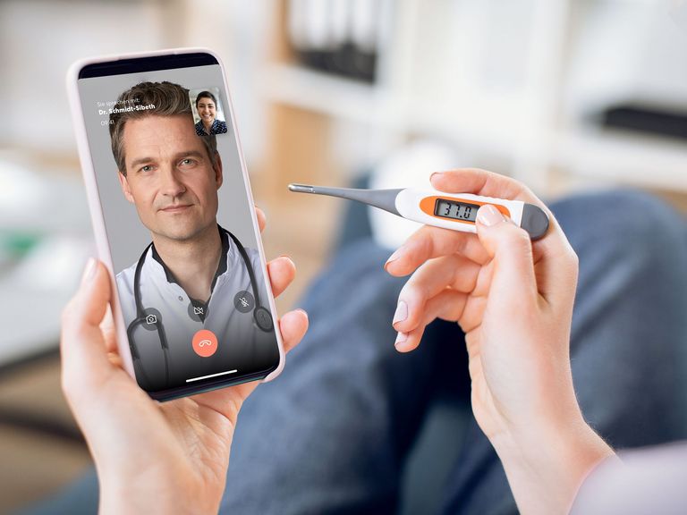 Hände halten ein Fieberthermometer und ein Smartphone, auf dem ein Videochat mit einem Arzt zu sehen ist
