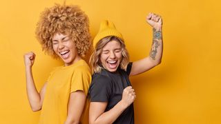Fusionsjubel Zwei Frauen jubeln mit erhobenen Händen vor gelbem Hintergrund