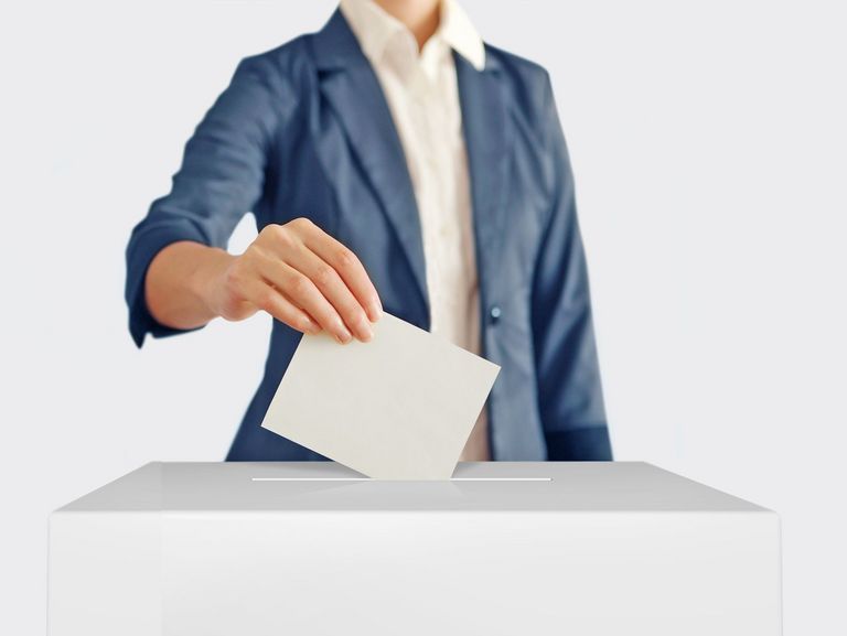 Frau in einem dunkelblauen Jacket steckt ihren Wahlzettel in eine helle Wahlbox