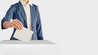 Frau in einem dunkelblauen Jacket steckt ihren Wahlzettel in eine helle Wahlbox