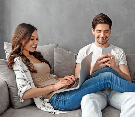 Pärchen sitzt fröhlich mit Smartphone und Laptop auf einem grauen Sofa