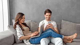 Pärchen sitzt fröhlich mit Smartphone und Laptop auf einem grauen Sofa