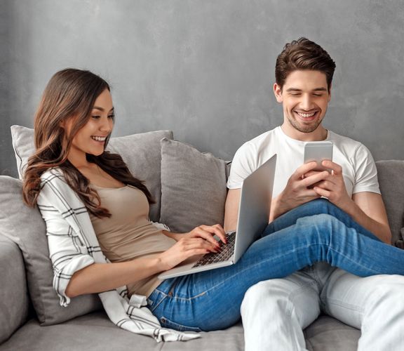 Pärchen sitzt fröhlich mit Smartphone und Laptop auf einem grauen Sofa