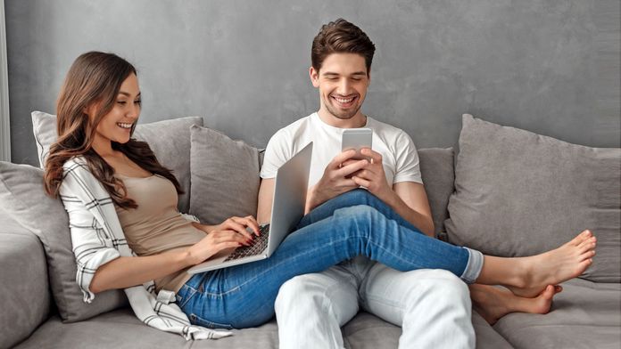 Pärchen sitzt fröhlich mit Smartphone und Laptop auf einem grauen Sofa