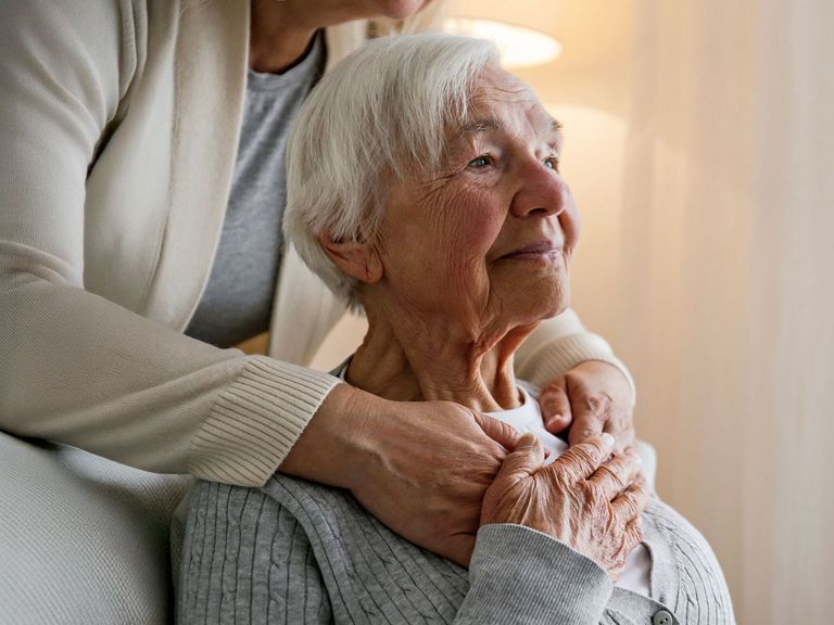 Frau betreut eine Pflegebedürftige und legt ihre Arme um deren Schultern