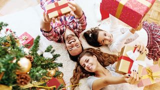 Familie liegt fröhlich unter einem Weihnachtsbaum und streckt Geschenke in die Höhe in Richtung Kamera