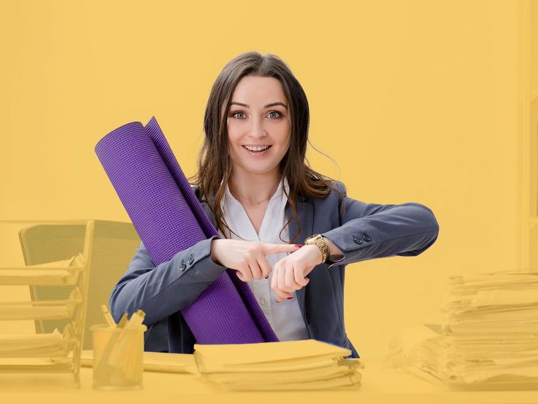 Azubi sitz an einem vollem Schreibtisch mit Yogamatte und zeigt auf die Uhr	