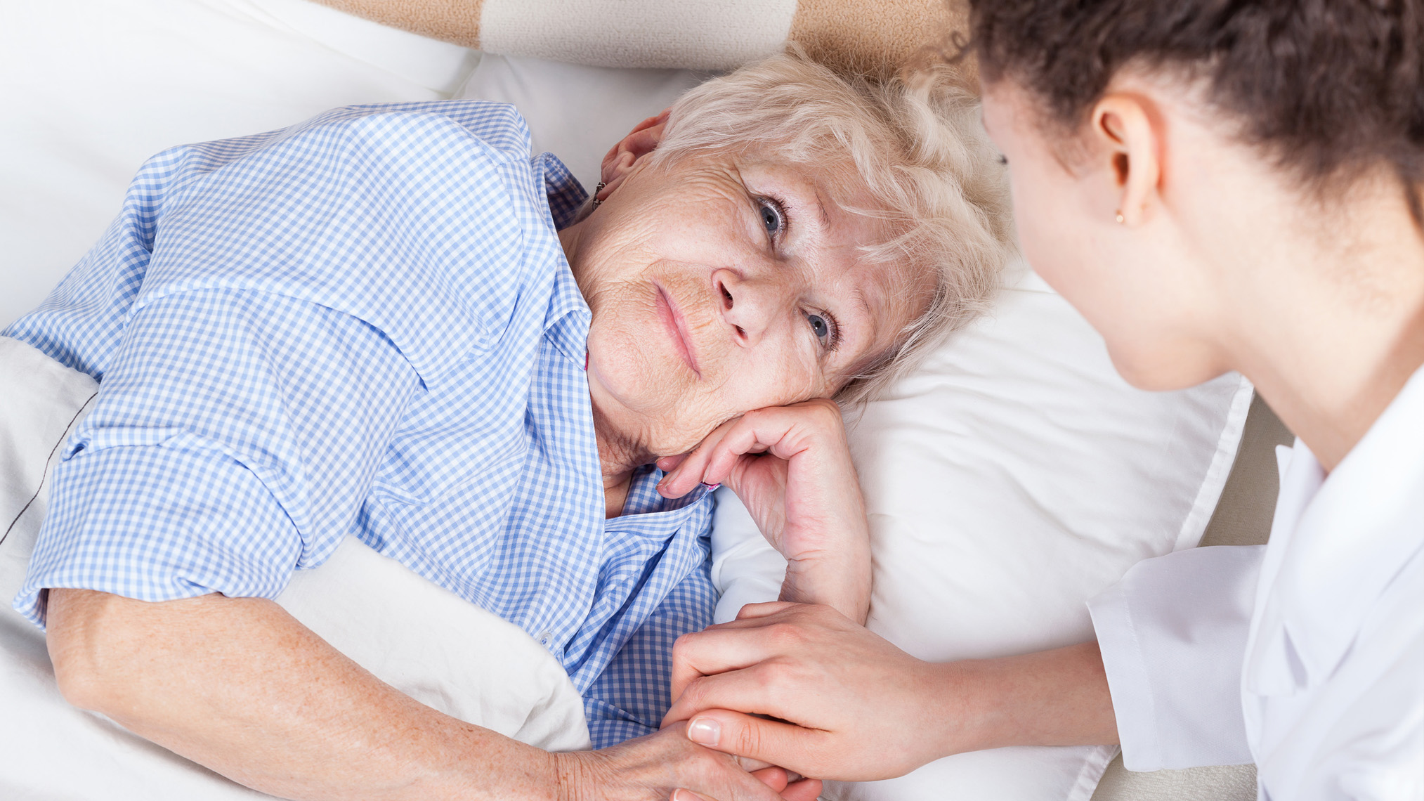 Ältere Frau im Bett wird von junger Pflegerin versorgt