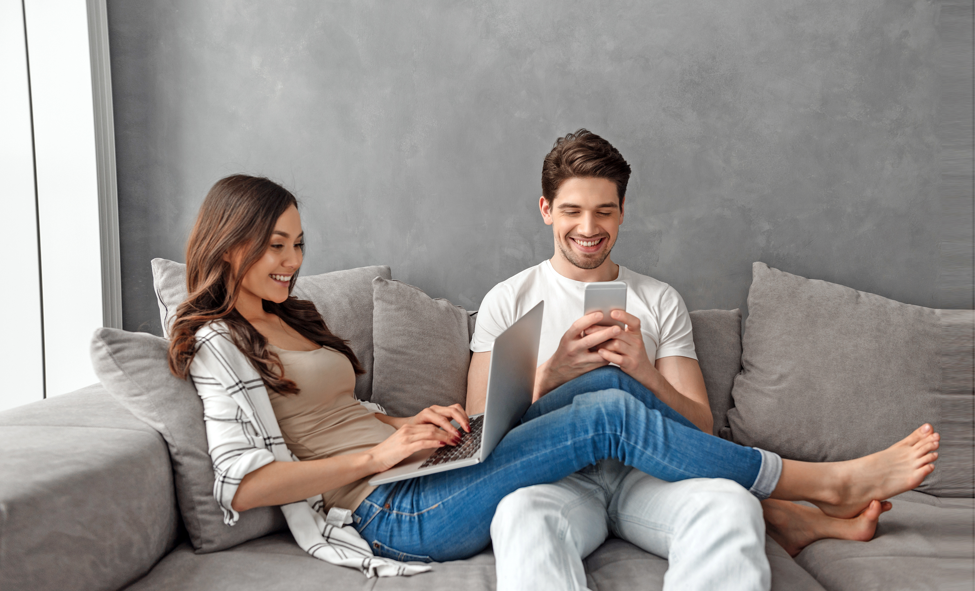 Pärchen sitzt fröhlich mit Smartphone und Laptop auf einem grauen Sofa