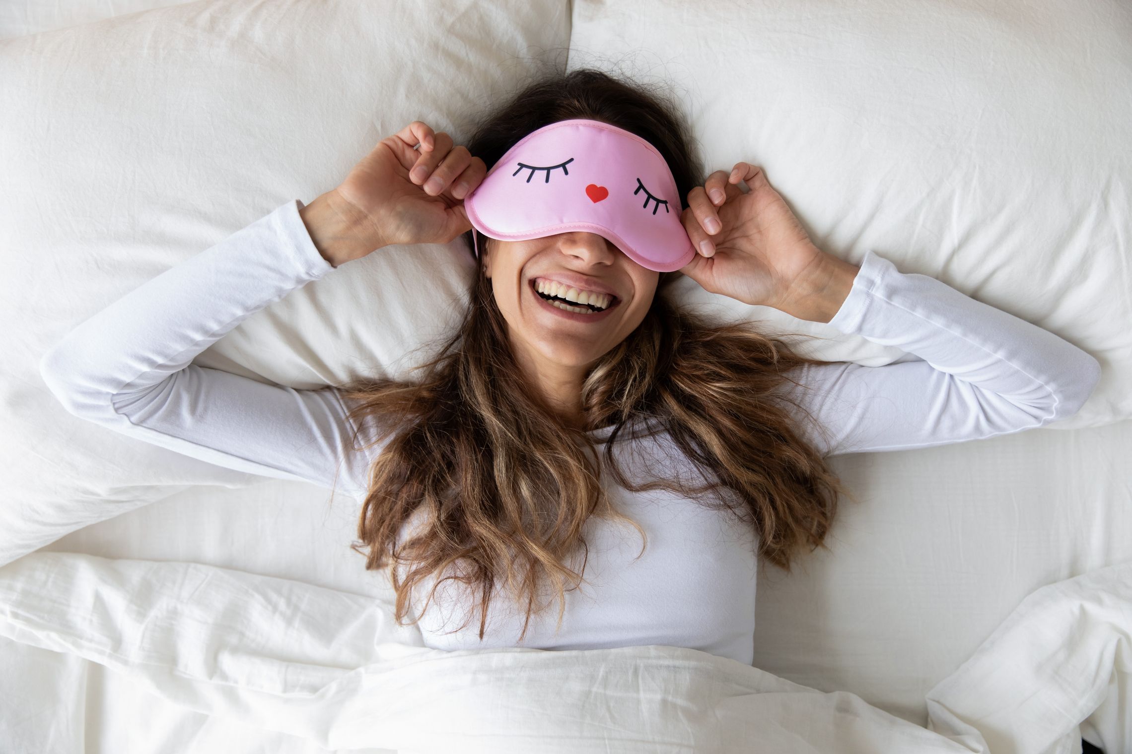 Frau mit einer pinken Schlafmaske liegt glücklich auf einem Bett mit weißem Bettzeug