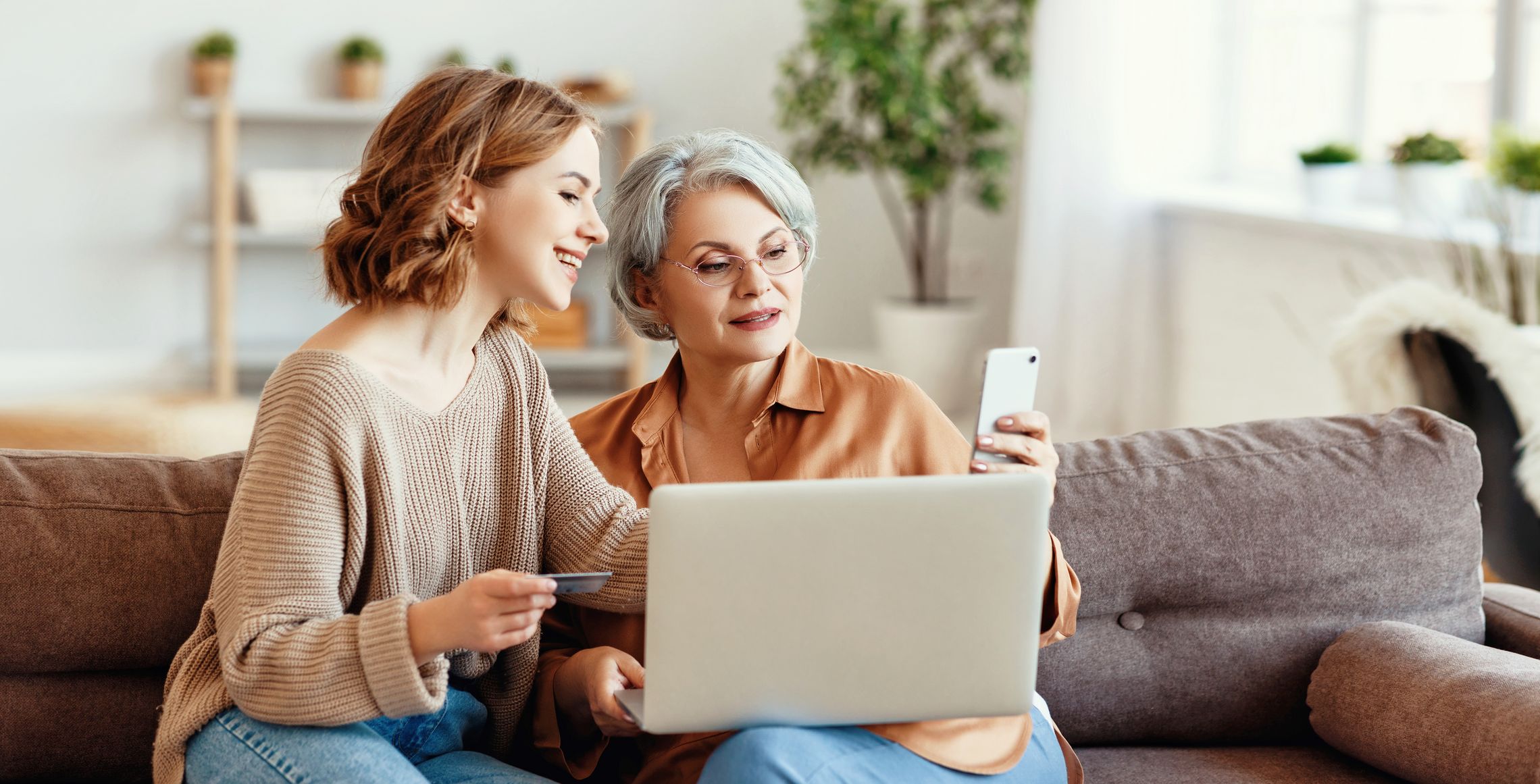 Zwei Frauen sitzen im Wohnzimmer auf einem Sofa und nutzen digitale Angebote per Laptop und Smartphone