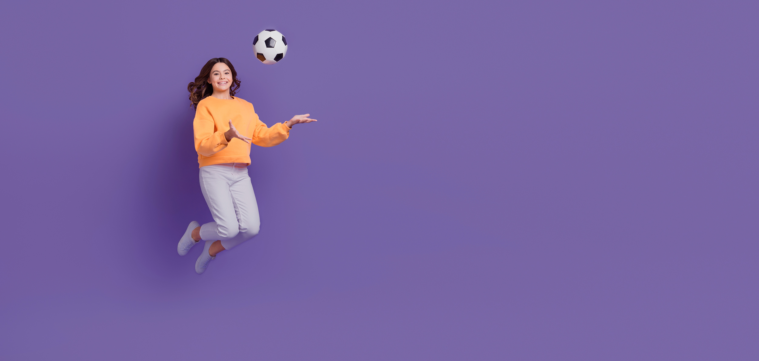 Mädchen im orangefarbenen Pullover springt mit einem Fußball in der Hand und vor einem lilafarbenen Hintergrund in die Luft