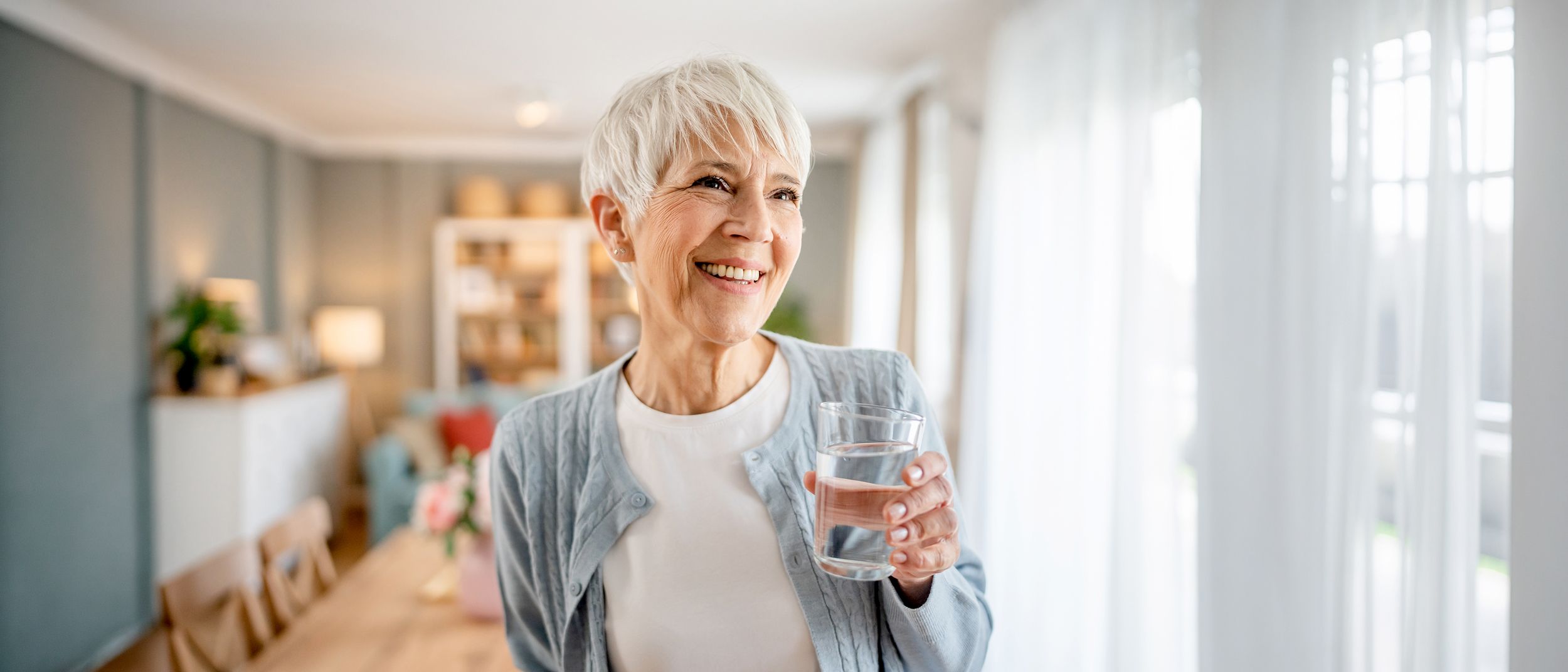 Frau lächelnd im Wohnzimmer mit einem Glas Wasser in der Hand