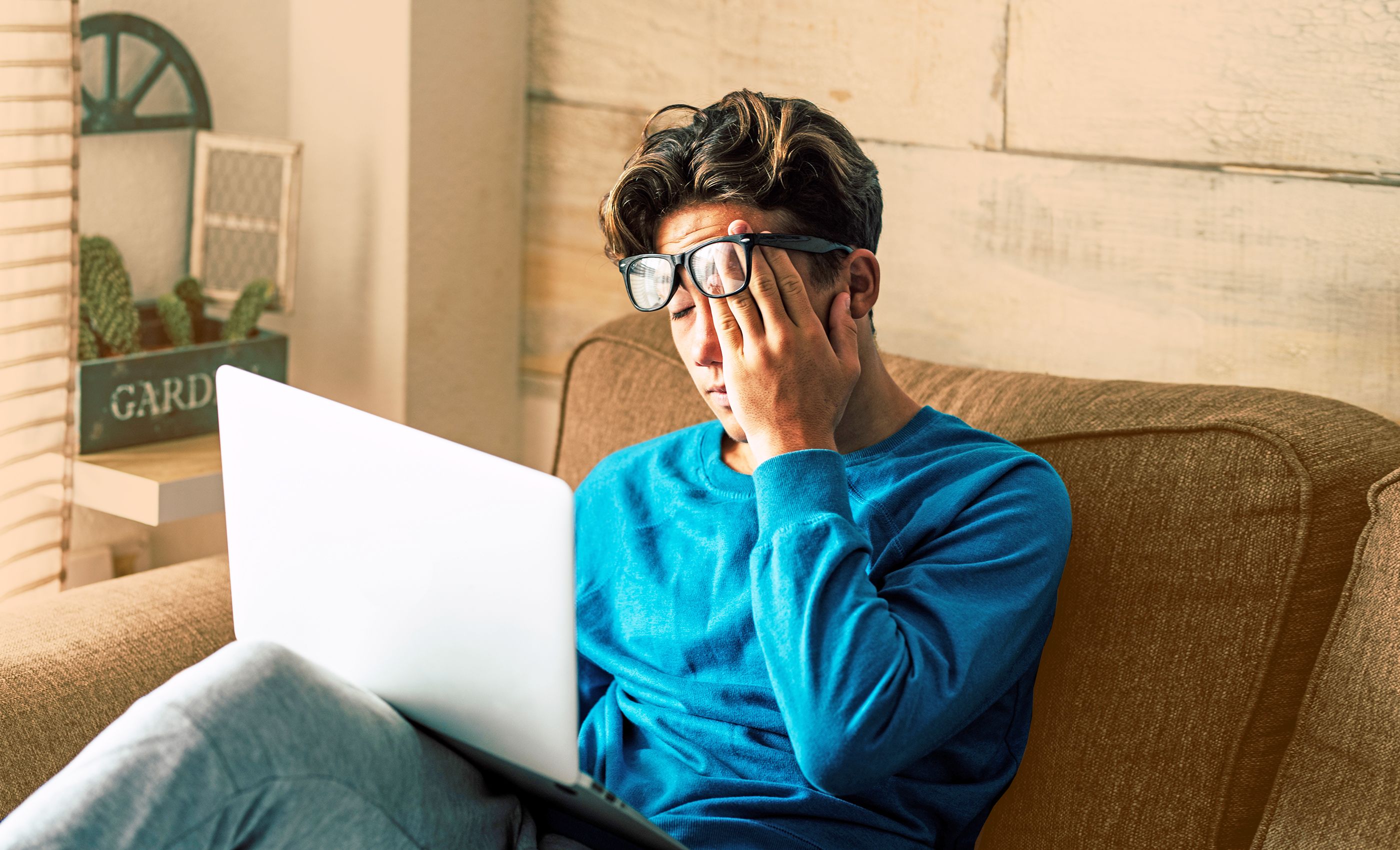 Jugendlicher sitzt auf der Couch, hat einen Laptop auf dem Schoß und reib sich mit einhändig die Augen und verschiebt dabei seine Brille