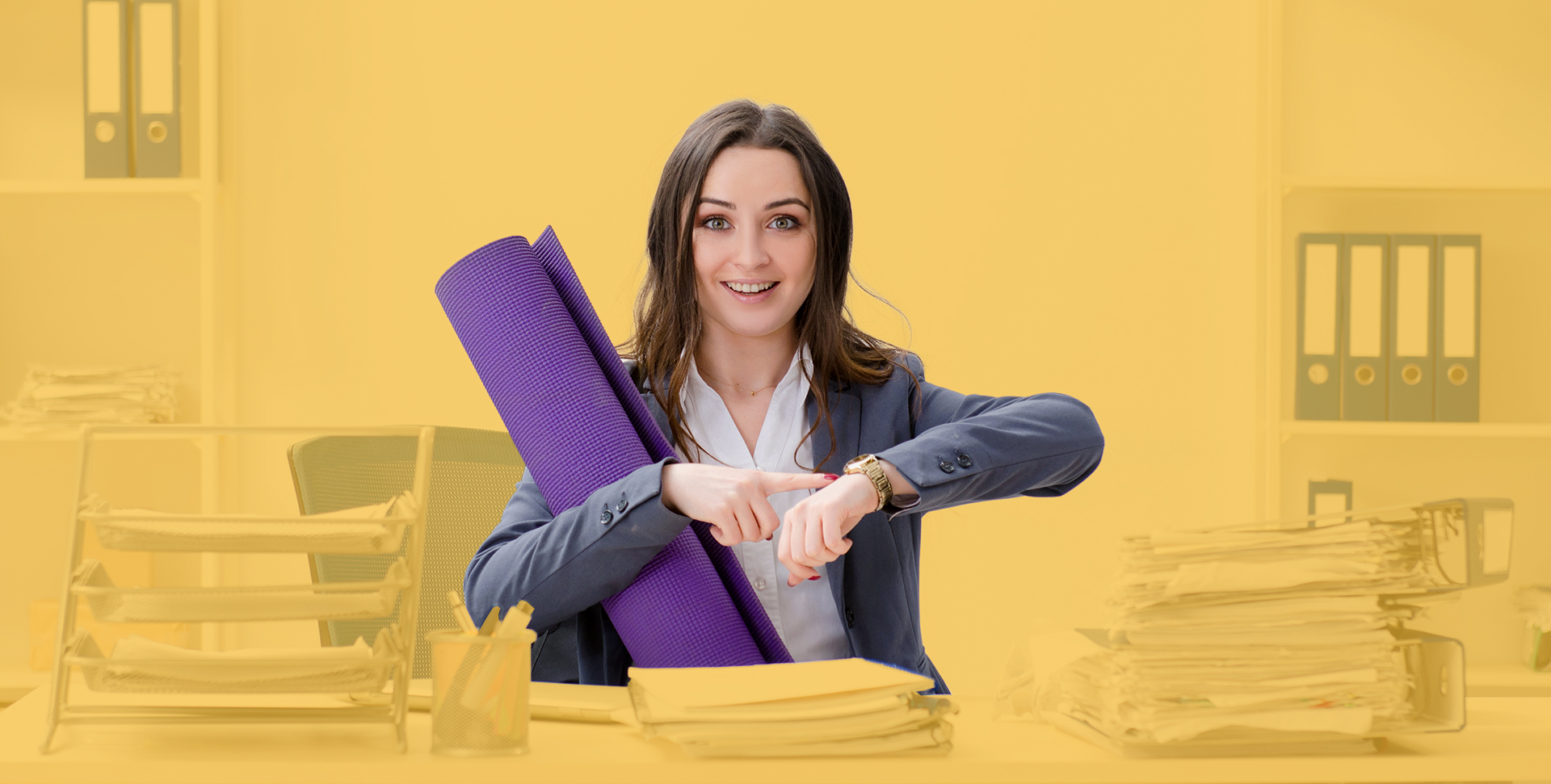 Azubi sitz an einem vollem Schreibtisch mit Yogamatte und zeigt auf die Uhr	