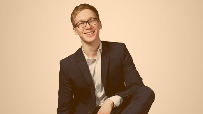 Jugendlicher, mit Brille und gepflegtem Haar, sitzt auf einem Stuhl und trägt einen blauen Anzug mit weißem Hemd darunter 