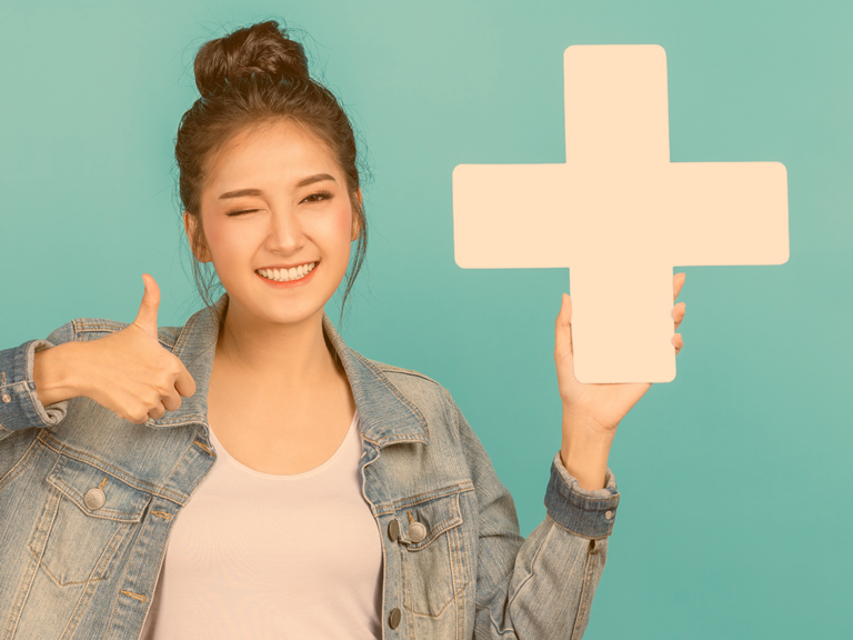 Junge Frau mit Dutt steht vor farbiger Wand und hält in der einen Hand ein großes, weißes  Pluszeichen und zeigt mit der anderen Hand Daumen hoch