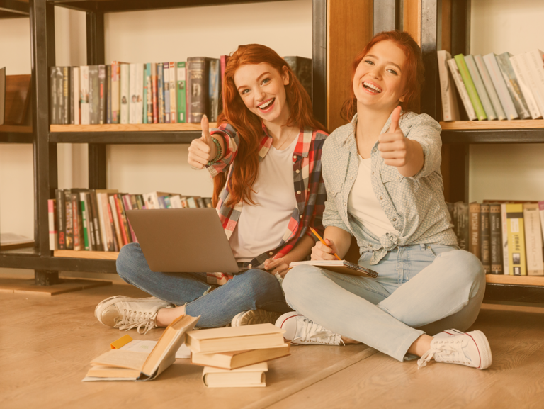 Zwei Jugendliche sitzen vor einem Bücherregal auf dem Boden und halten eine Daumen hoch