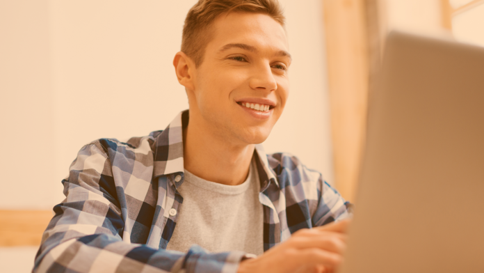 Blonder Jugendlicher im blaukariertem Hemd am Schreibtisch vor Laptop sitzend