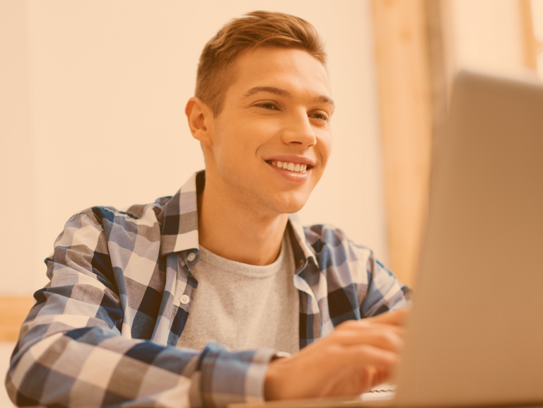 Blonder Jugendlicher im blaukariertem Hemd am Schreibtisch vor Laptop sitzend